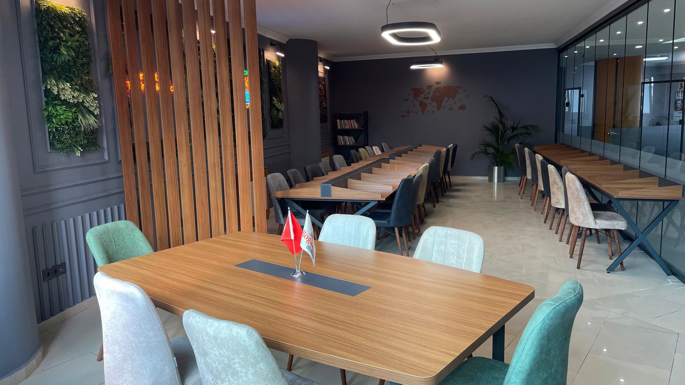 Uzun ahşap çalışma masaları, bitki duvarı ve cam bölmeli etüt salonu.