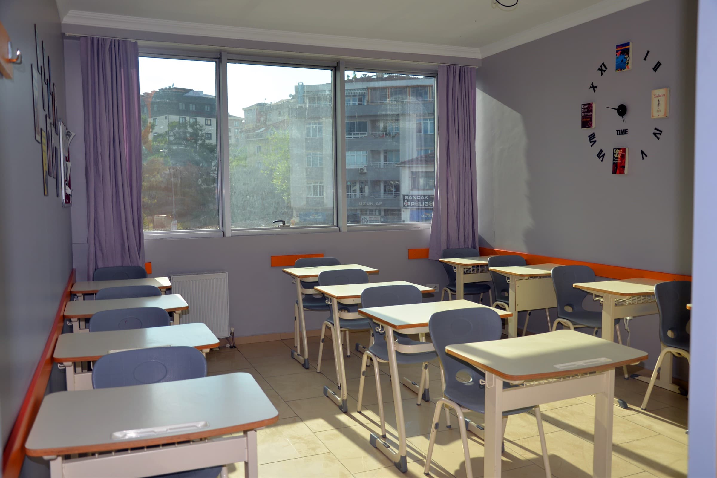 Pencereden şehir manzarası alan boş bir derslik ve tek kişilik sıralar.